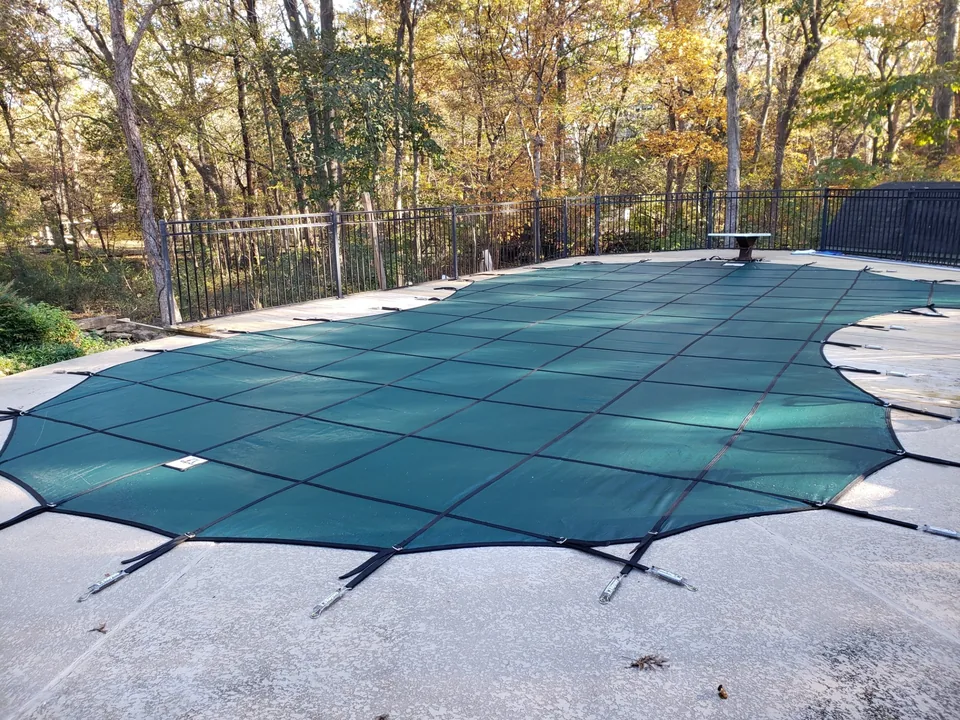 Green mesh pool cover following the pool’s curved outline, with straps secured to deck anchors