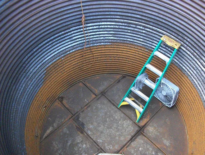 Top-down view of a bare corrugated metal tank interior with a seamed steel floor, a ladder, and a fan